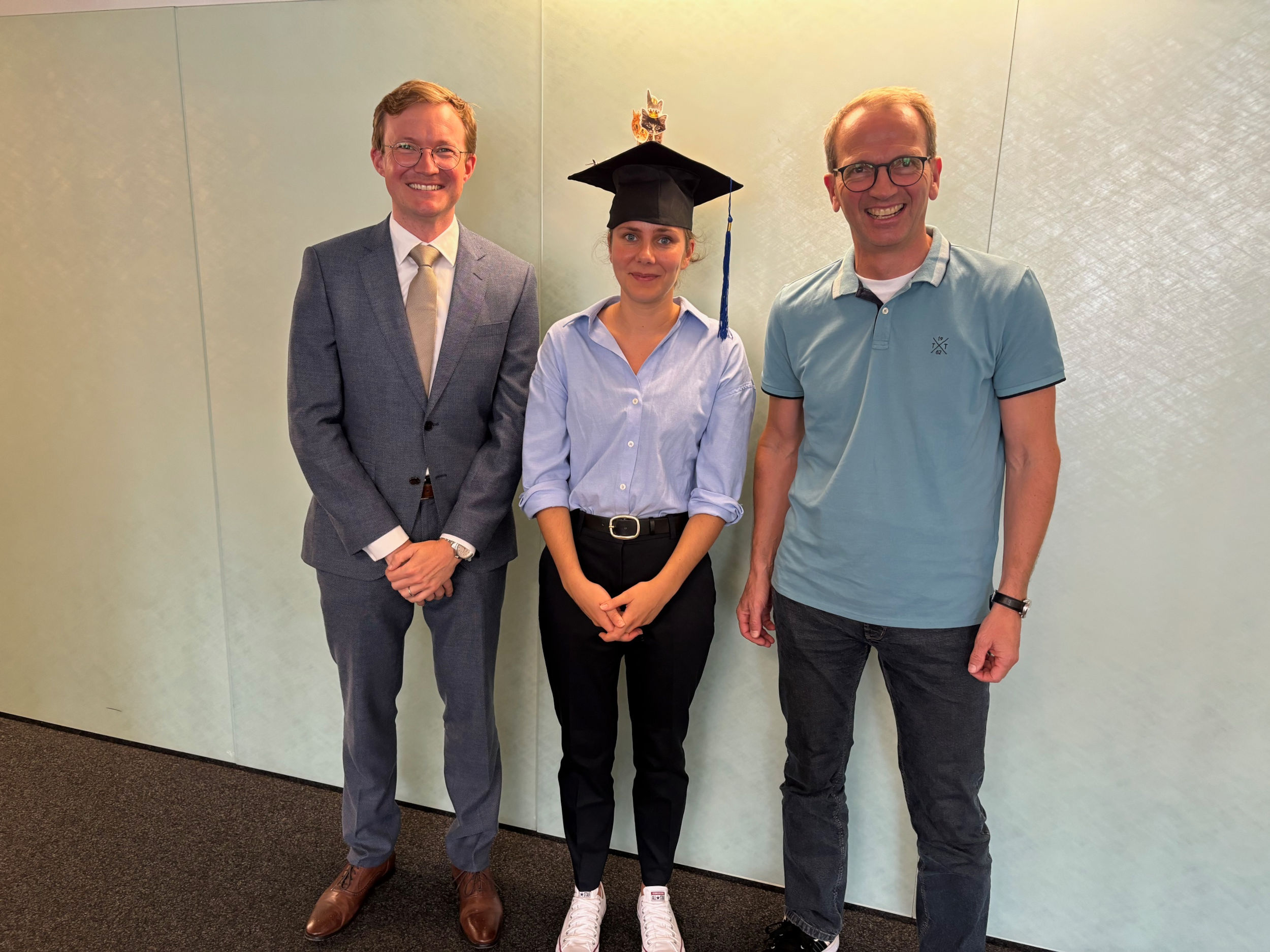 Sarah, Prof. Wagner, and Dr. Winkenbach standing together and smiling. Sarah wears an academic hat typical for graduations