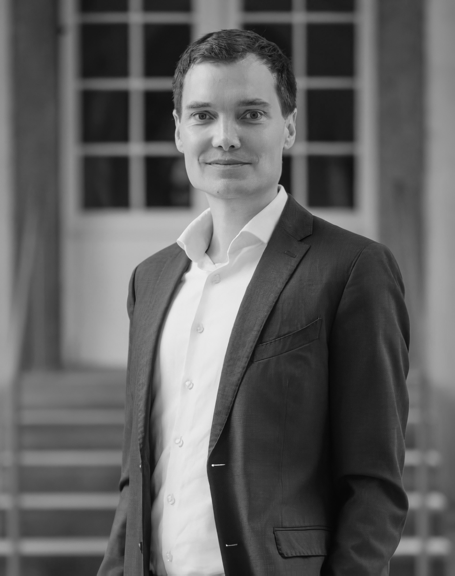 Prof. Durach in a suit, smiling. Standing in front of some chairs and a wood and glass door.